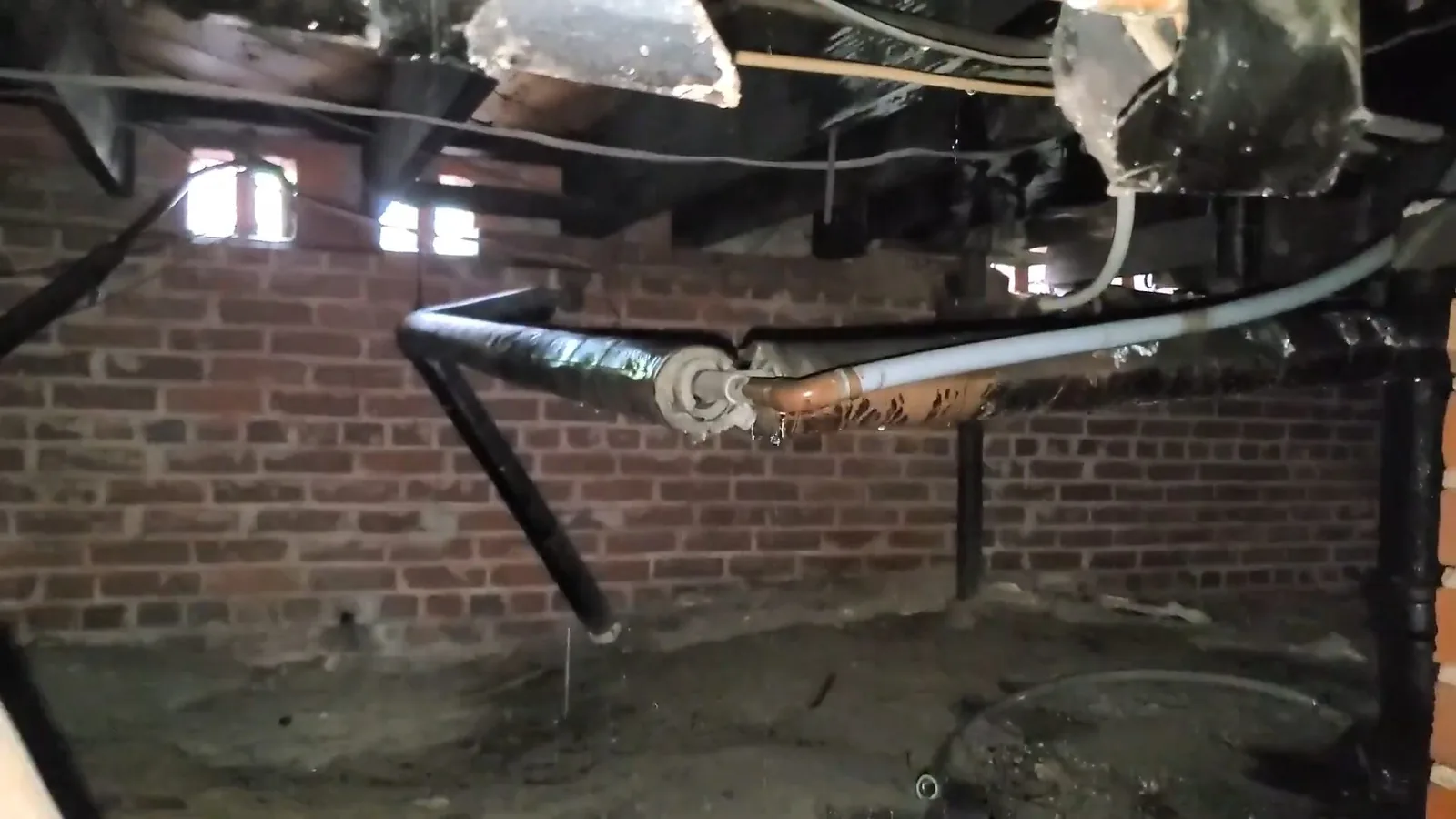 Black drain pipe and a copper line under floor joists along a brick wall.