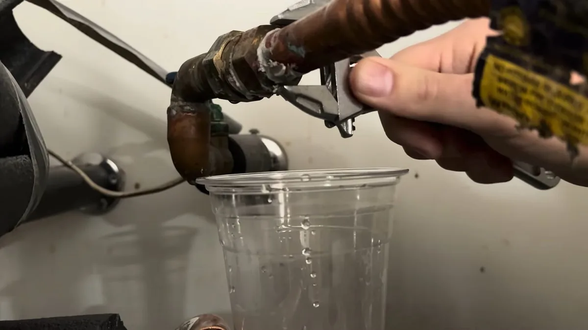 Wrench loosening a drain fitting on a water heater, with water running into a clear plastic cup.
