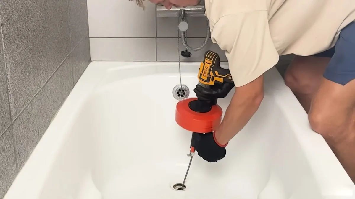 Person using a drill-powered drain auger with a coiled cable in a white bathtub drain.