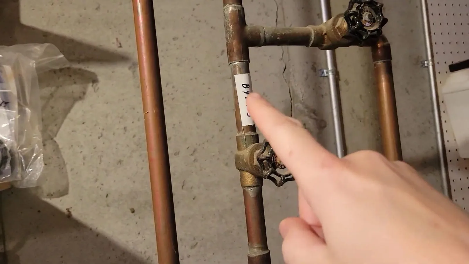 Finger pointing to a labeled valve on copper supply pipes along a basement wall.
