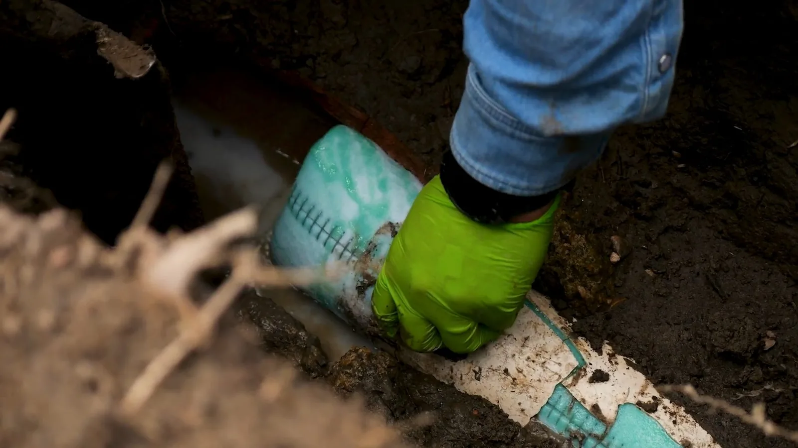 Gloved hand guiding a green patterned liner into a pipe in a dug trench.