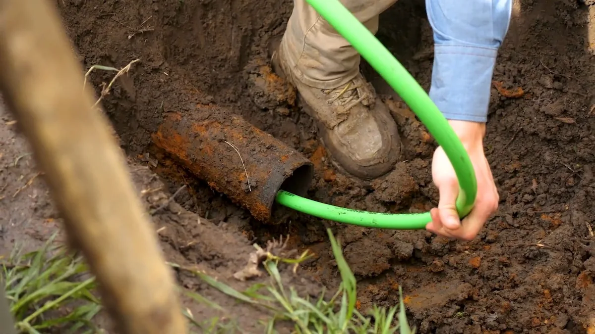 Green hose fed into the end of a rusted pipe in an open trench, with a boot beside it.