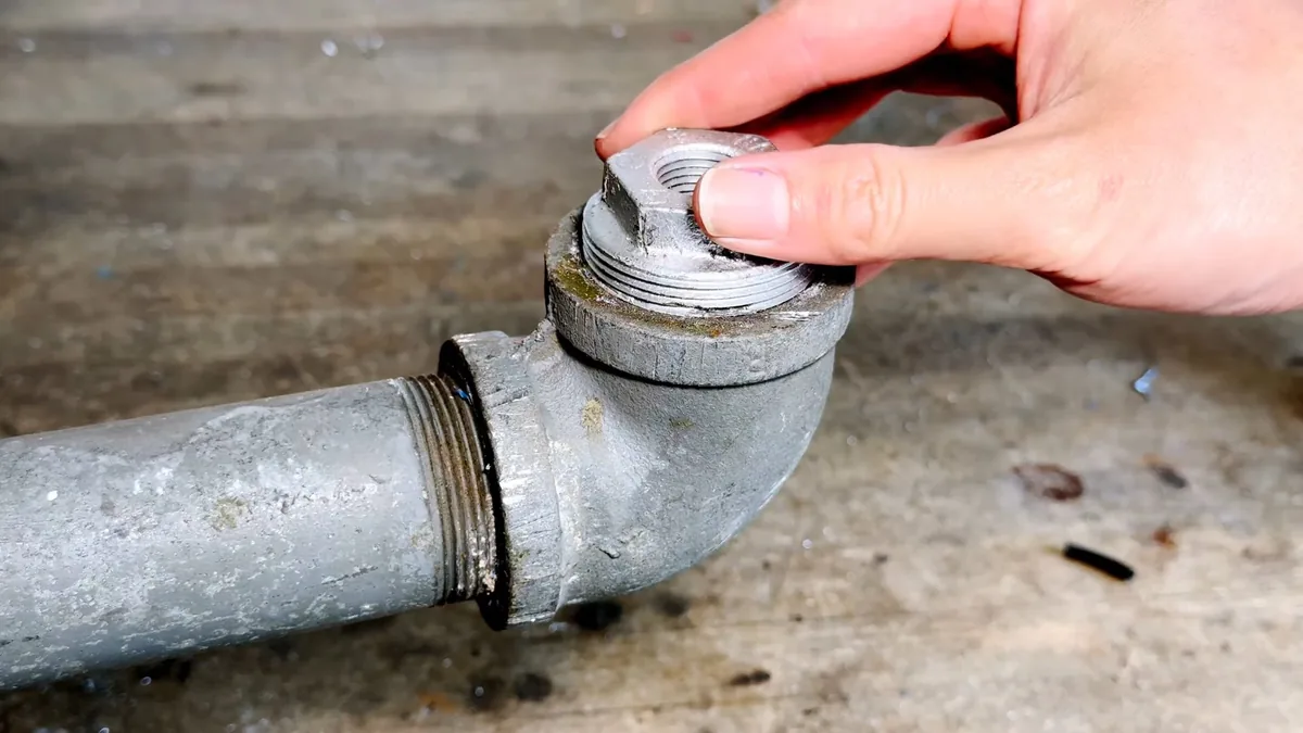 Hand holding a threaded plug above a galvanized cleanout fitting on a pipe.