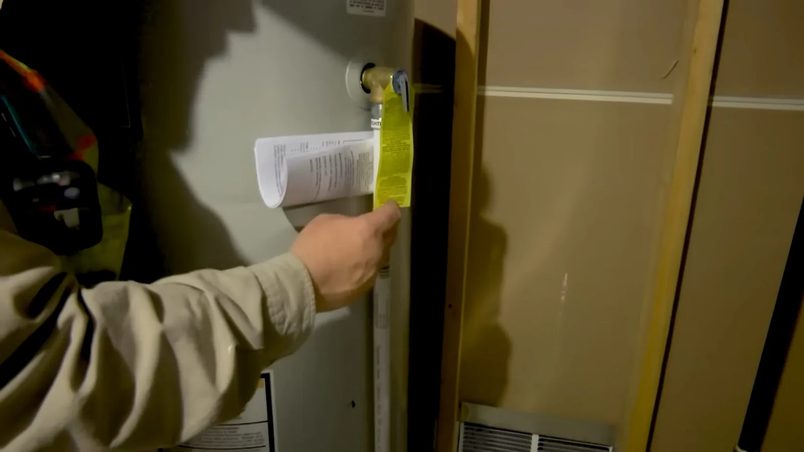 Hand holding a yellow tag and papers at a valve beside a water heater cabinet.