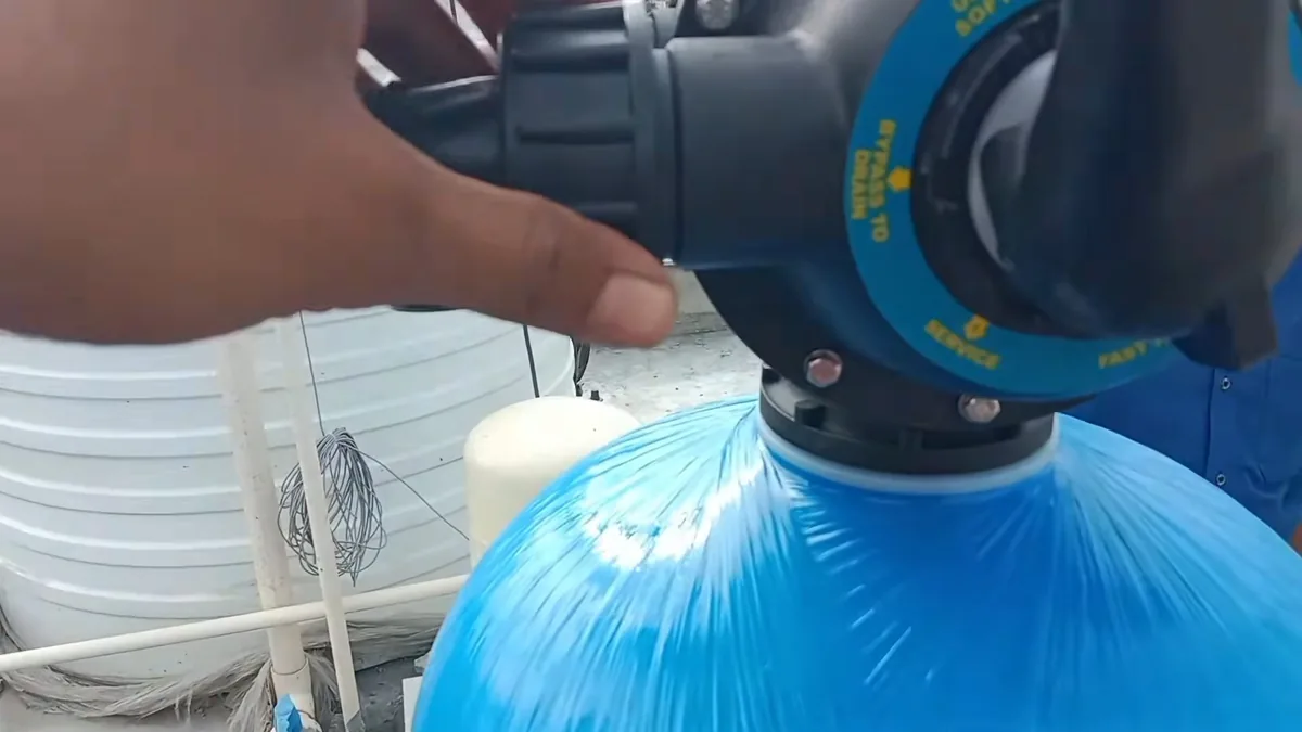 Hand on the control head of a blue softener tank, marked with bypass and service positions.