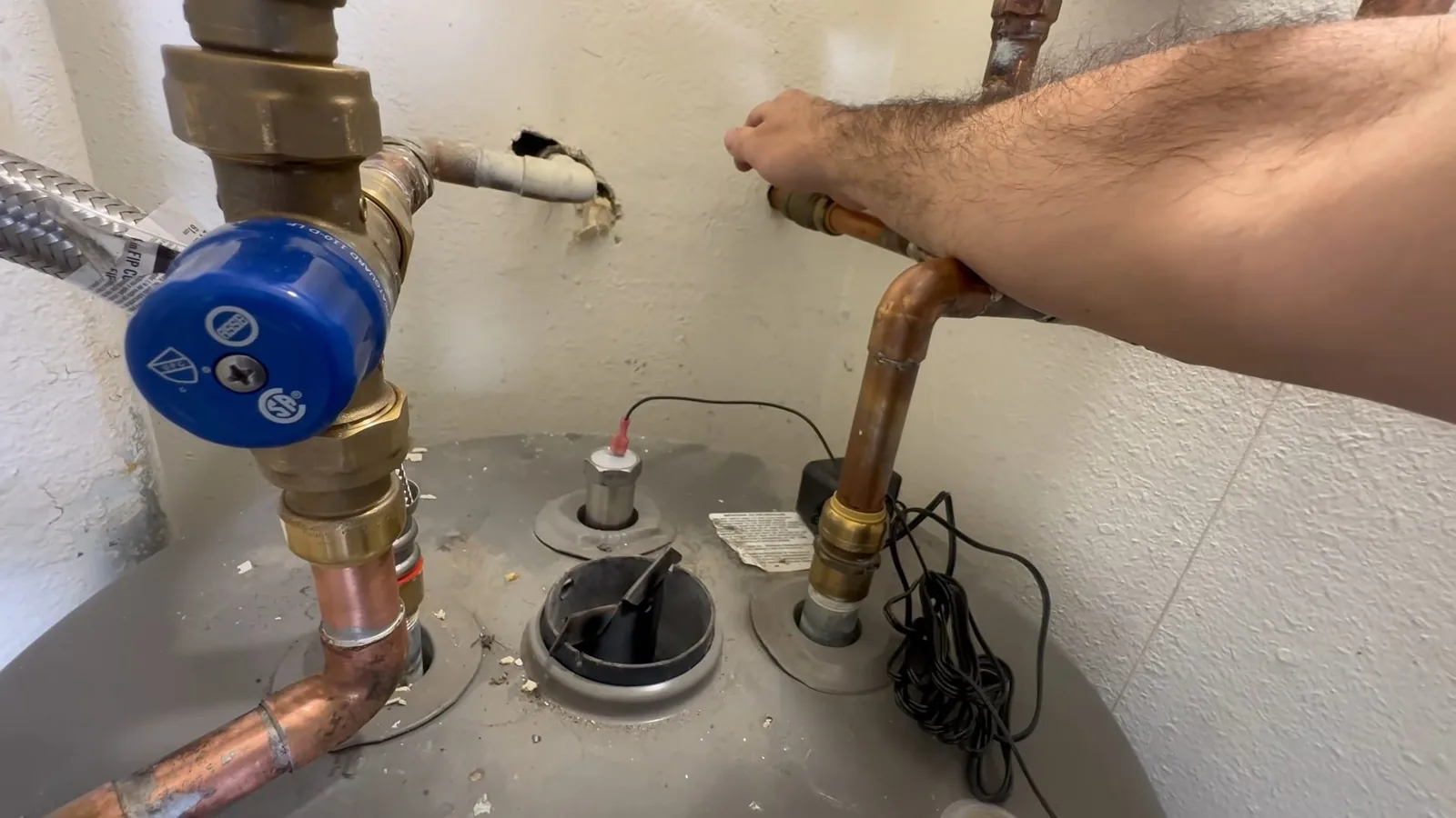 Hand on a copper supply pipe at the top of a tank water heater, beside a blue-handled valve and a power cable.
