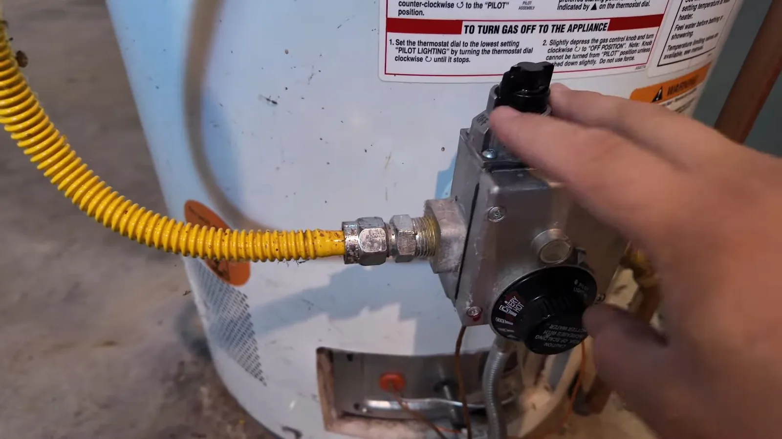 Hand turning the gas control knob on a water heater with a yellow flexible gas line attached.