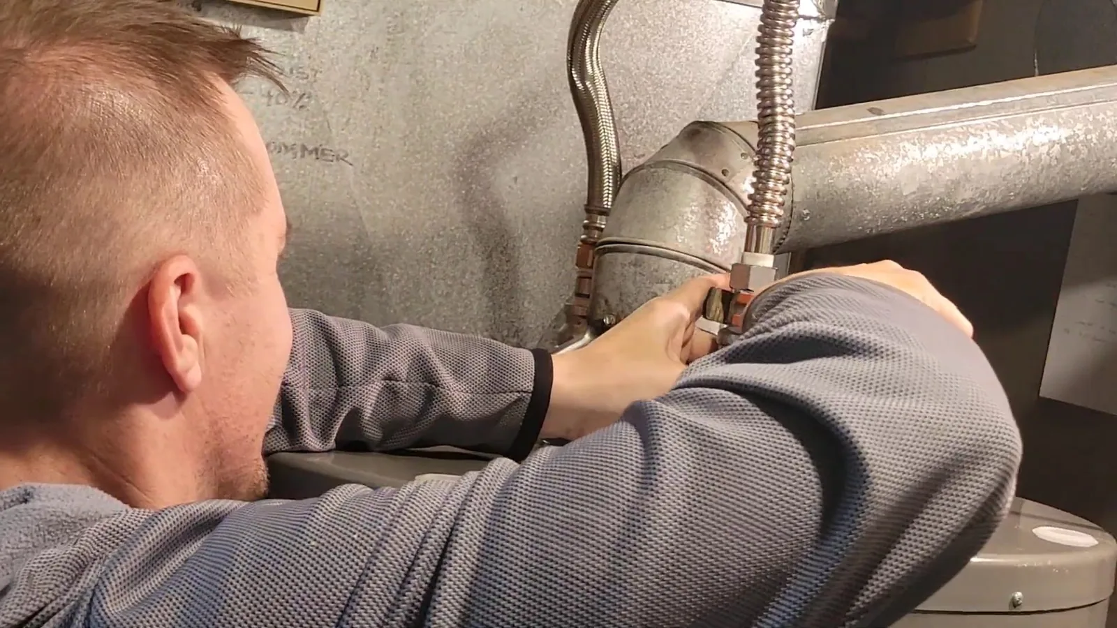 Man reaching to flexible gas lines and a duct at a water heater in a basement.