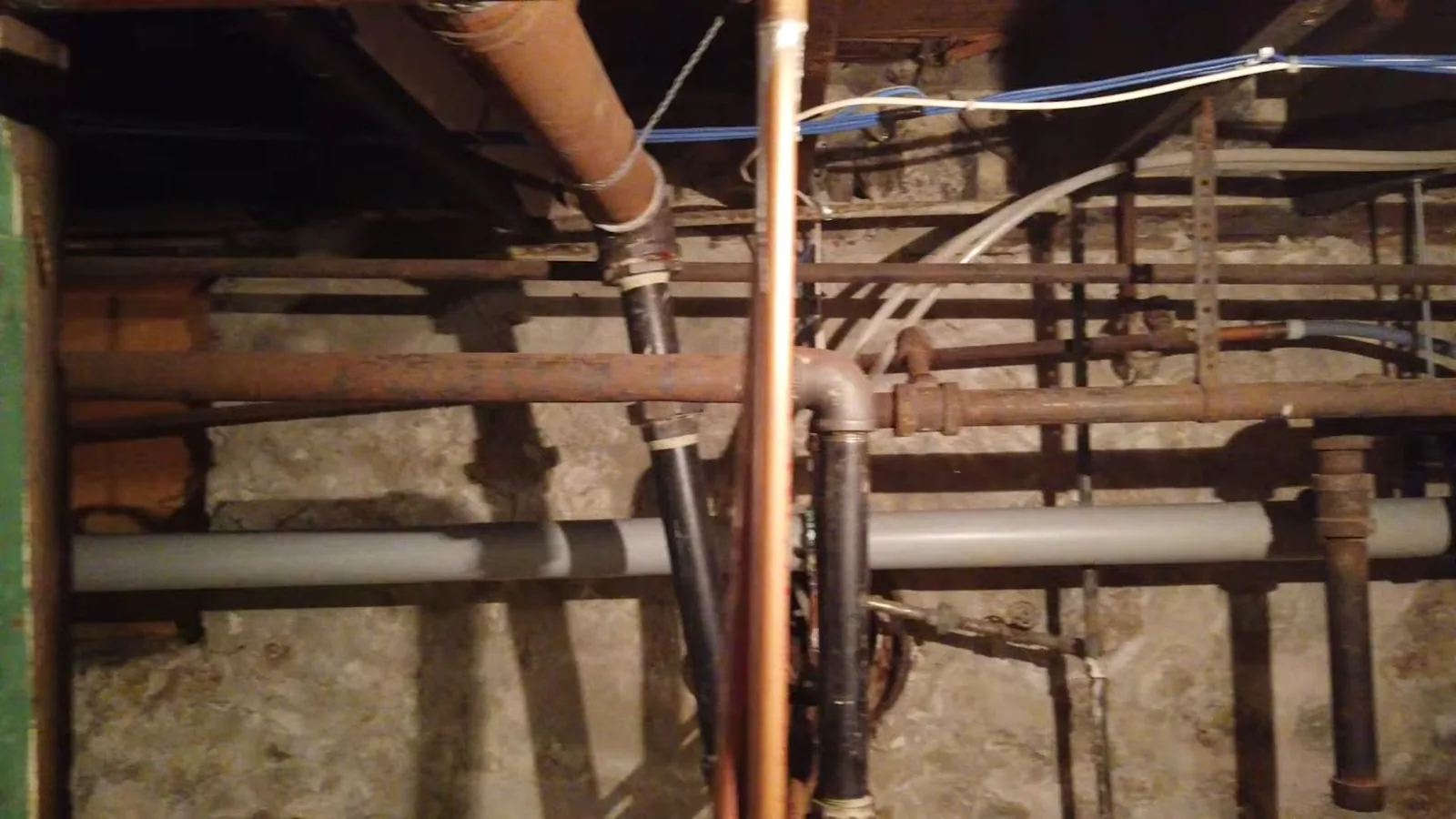 Rusted iron, black drain and copper pipes running under basement floor joists.