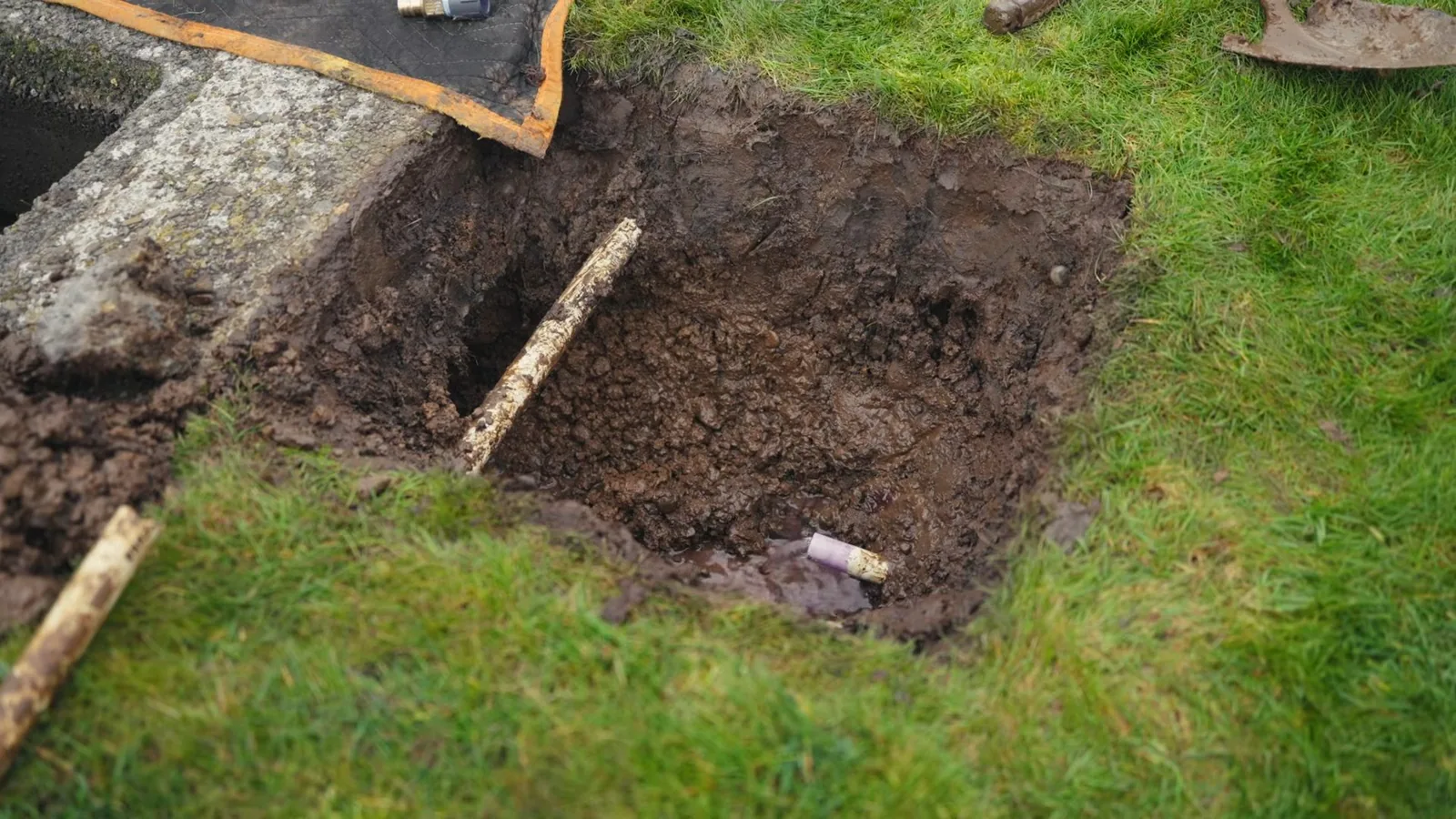 Square hole dug in a lawn beside a concrete box, with a shovel handle at the edge and a short pipe at the bottom.