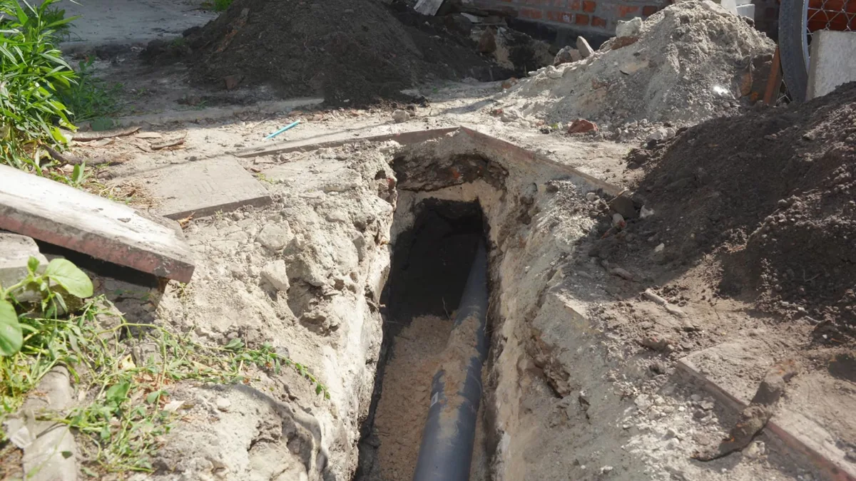 Narrow trench in a yard exposing a dark sewer pipe, with piles of dirt beside it.