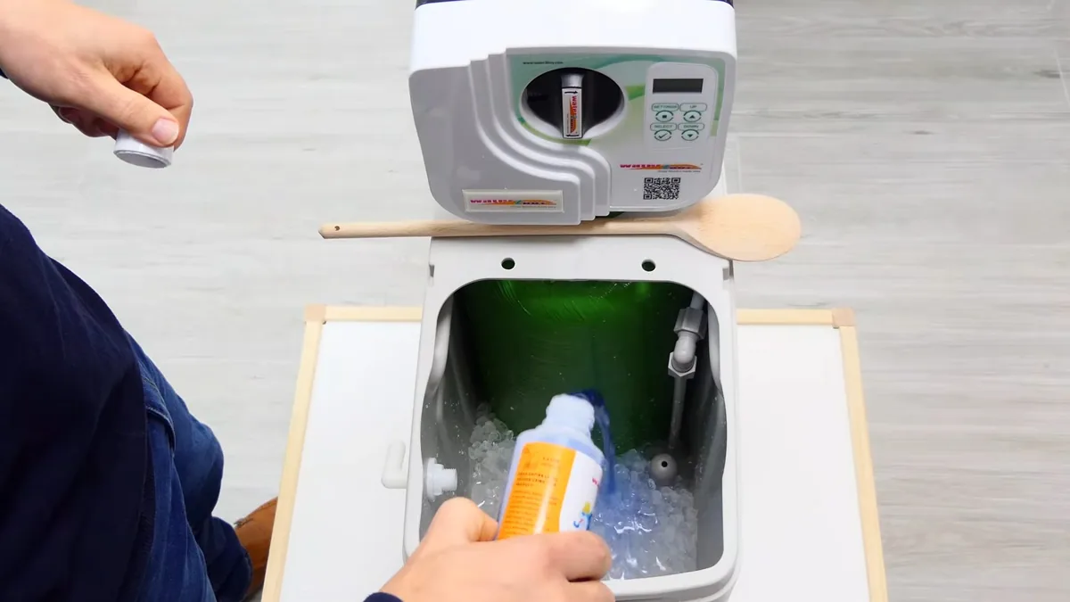 Open white water softener with a green tank inside and a hand adding product from an orange bottle.