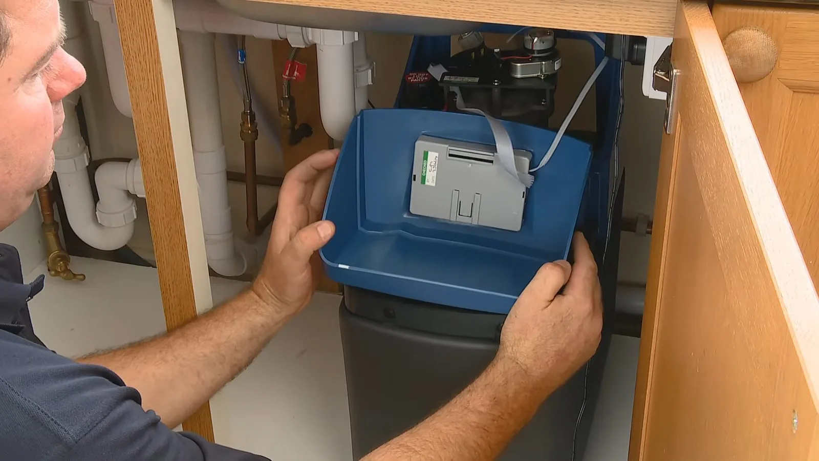 Plumber reaching into a kitchen sink cabinet to adjust a blue water softener unit.