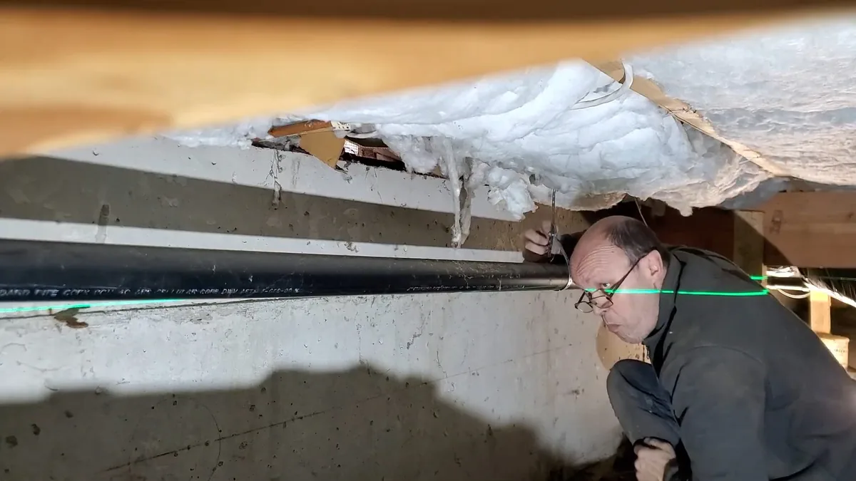 Plumber crouched in a crawlspace, looking along a pipe run under floor insulation with a green laser line across it.