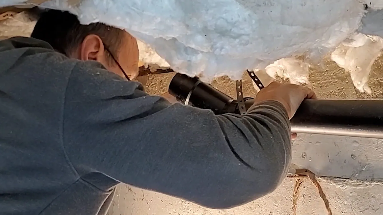 Man in a crawlspace reaching up to a black drain line under floor insulation.