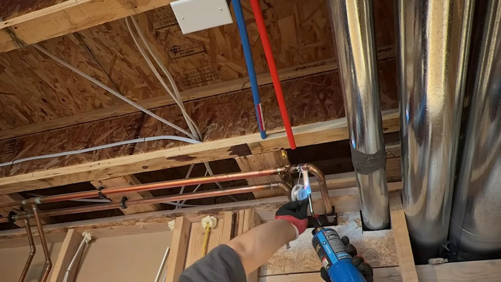 Plumber using a torch on a copper joint between basement joists, with new blue and red supply lines overhead.