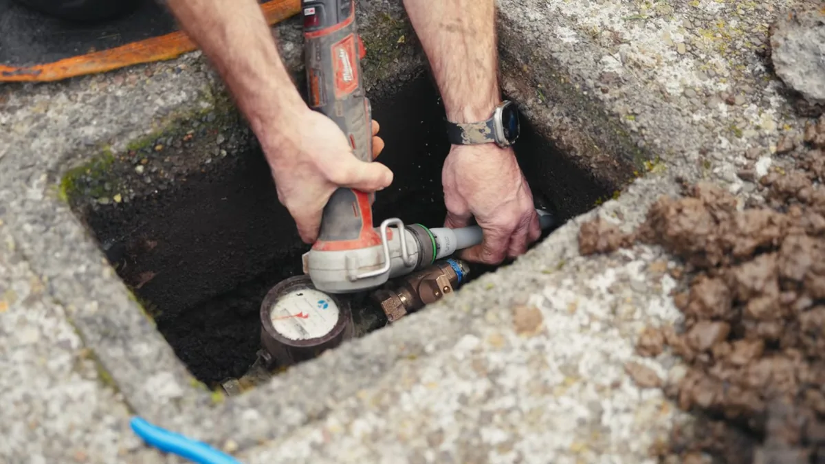 Plumber's hands using a power tool on a fitting beside a water meter inside a concrete ground box.