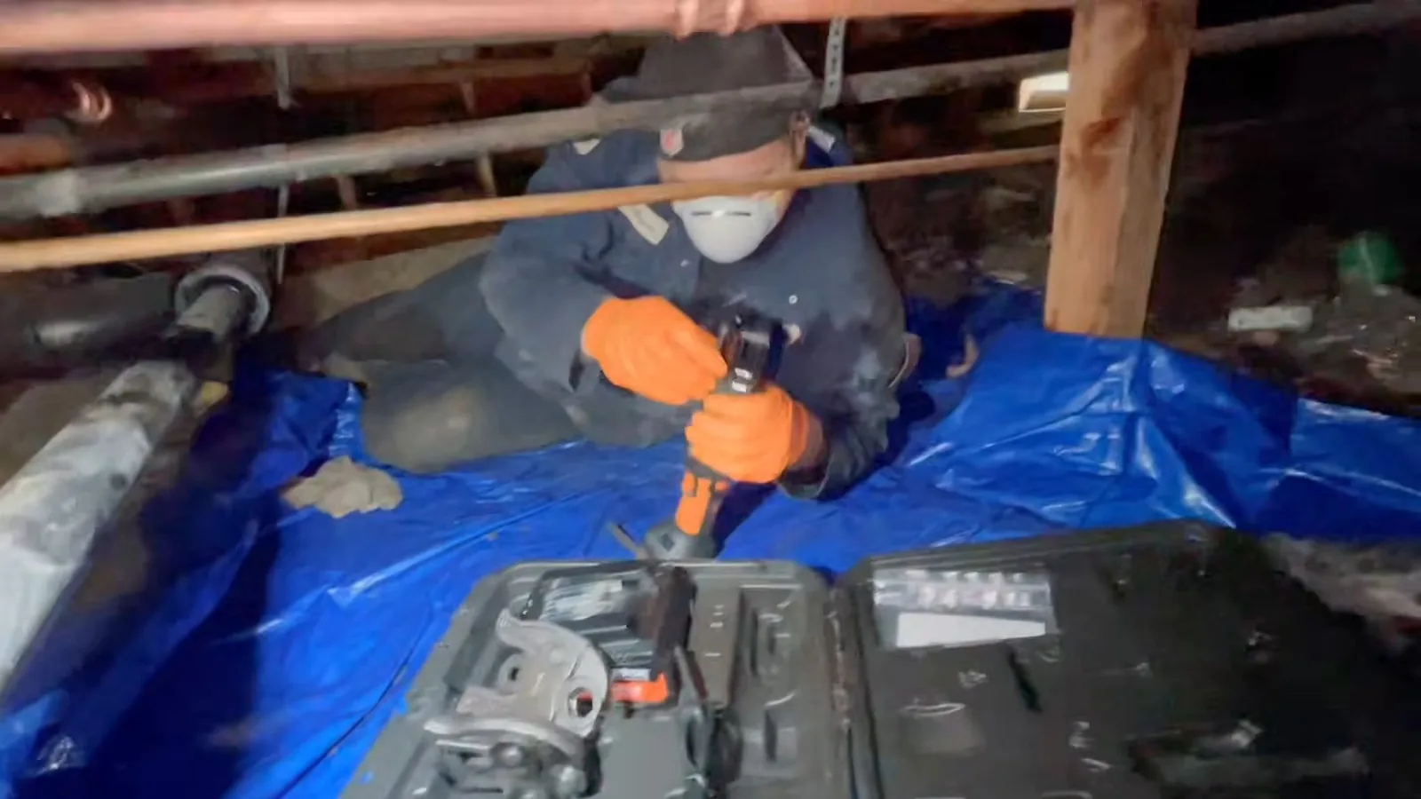 Plumber in a mask and orange gloves working on a pipe in a crawlspace over a blue tarp.