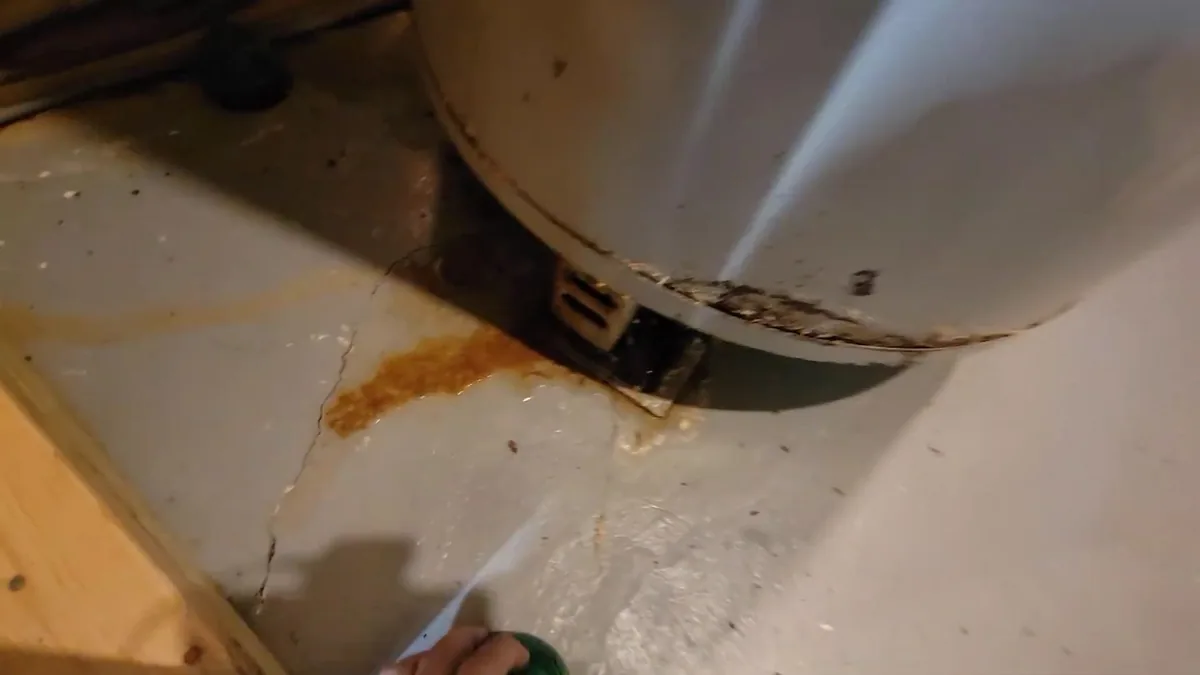 Rust stain and a wet streak on the floor below the base of a tank water heater.