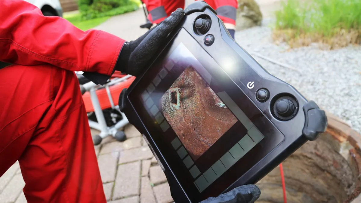 Gloved hands holding a sewer camera monitor showing a pipe interior, beside a brick-edged access opening.