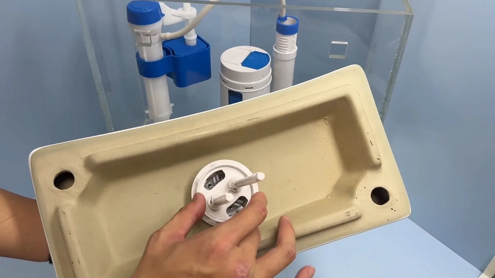 Hands holding a toilet tank lid, with its flush hardware underneath, in front of a clear tank holding a fill valve.