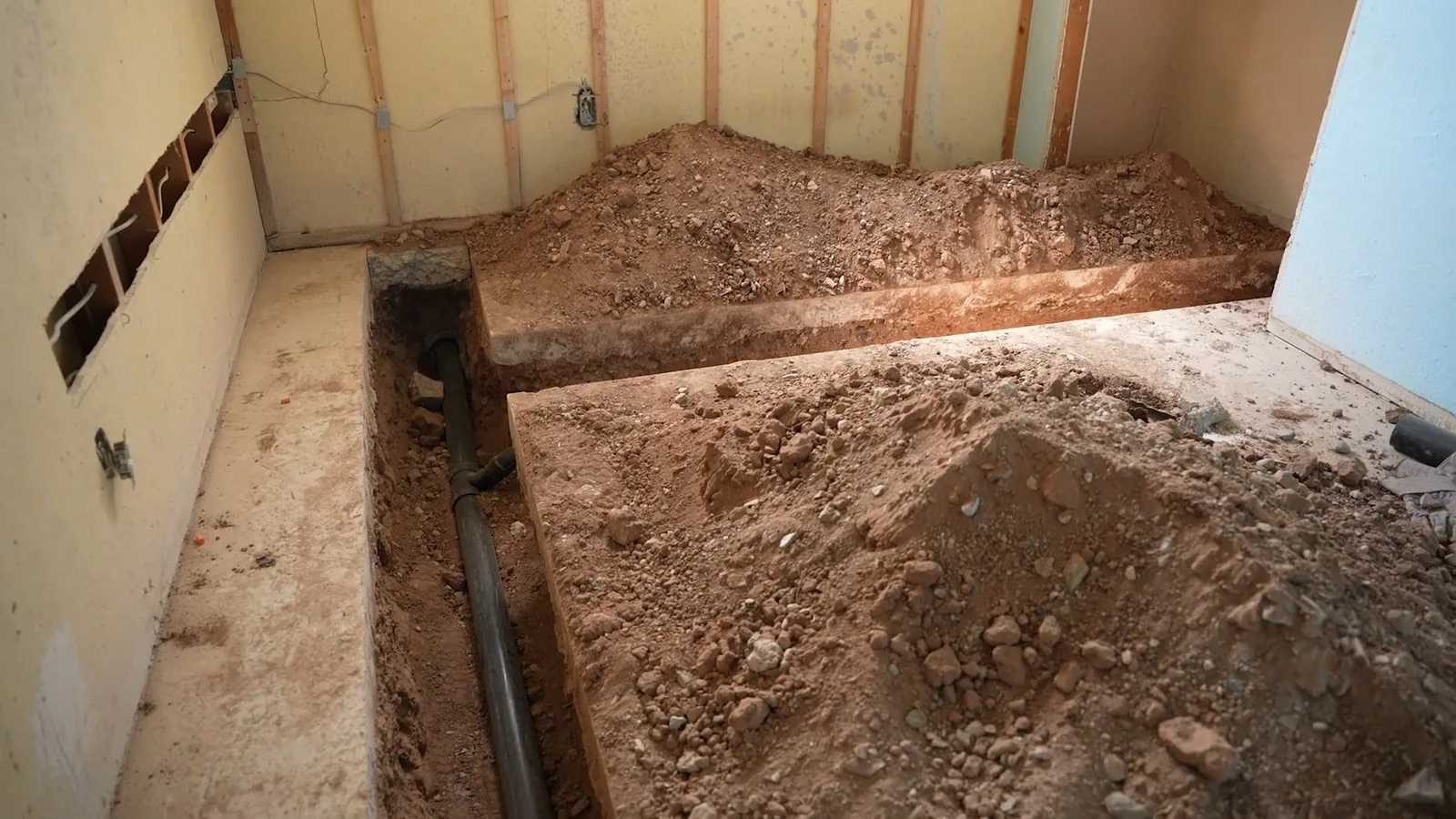 Trench cut along the base of a basement wall, with piles of soil on the floor and a pipe at the bottom.