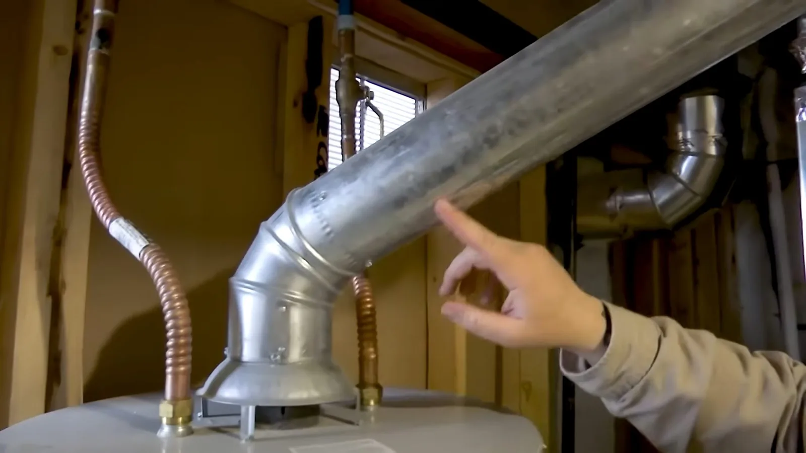Hand pointing to a galvanized vent connector on top of a tank water heater, with copper lines on either side.
