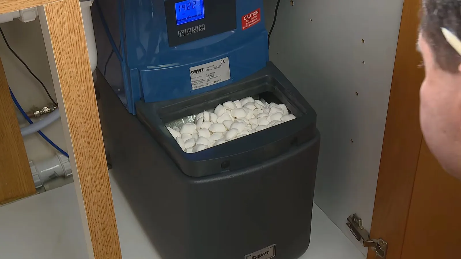 Blue water softener with its lid open, showing white salt tablets, set in a wooden cabinet.